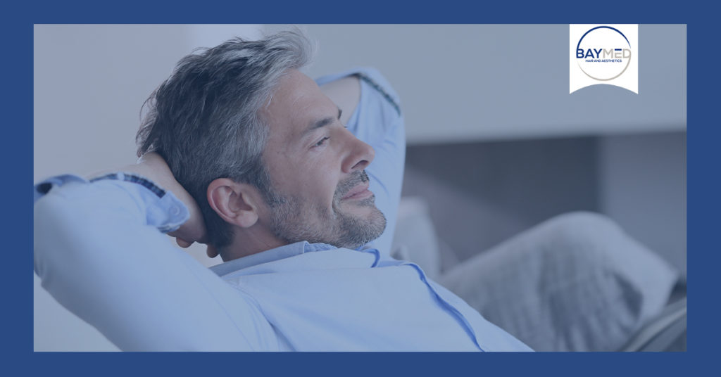 Man in blue button down relaxing with arms behind head.